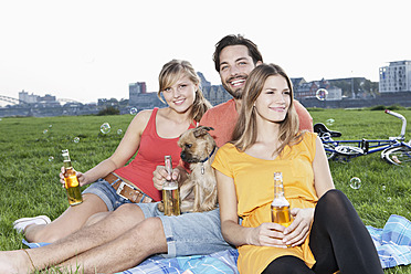 Deutschland, Köln, Junger Mann und Frau mit Hund und Bierflasche im Gras, lächelnd - FMKF000435