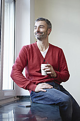 Deutschland, Köln, Älterer Mann sitzt mit Kaffee am Fenster, lächelnd - RHF000062