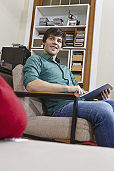 Germany, Cologne, Young man with digital tablet on chair, smiling - RHF000078