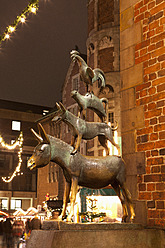 Germany, Bremen, Statue of Bremen City Minstrels in front of Christmas market at night - NDF000229