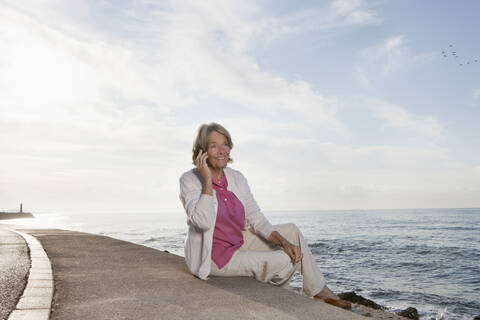 Spanien, Mallorca, Ältere Frau sitzt und benutzt Handy am Meer, lächelnd, lizenzfreies Stockfoto