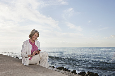 Spain, Mallorca, Senior woman sitting and using mobile phone at sea shore, smiling - SKF000772