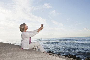 Spanien, Mallorca, Ältere Frau sitzt und benutzt Handy am Meer, lächelnd - SKF000773