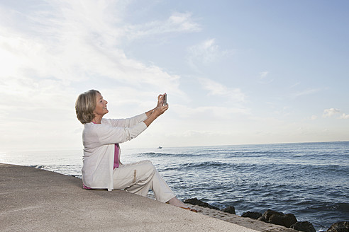 Spain, Mallorca, Senior woman sitting and using mobile phone at sea shore, smiling - SKF000773