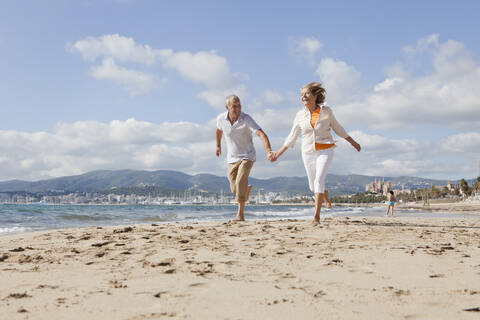 Spanien, Mallorca, Älteres Paar läuft am Strand entlang, lächelnd, lizenzfreies Stockfoto