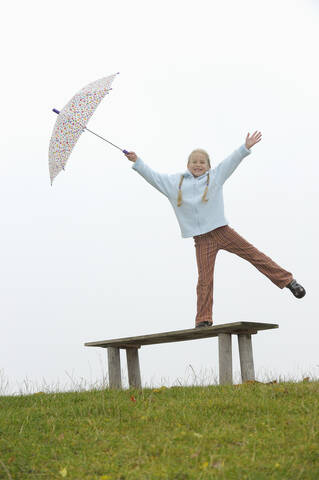 Deutschland, Bayern, Mädchen auf Bank stehend, lächelnd, Regenschirm haltend, lizenzfreies Stockfoto