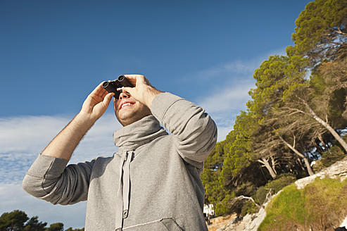 Spain, Mallorca, Young man looking through binocular, smiling - MFPF000019