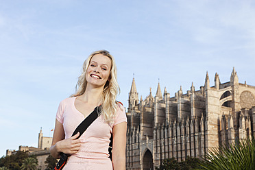 Spanien, Mallorca, Palma, Junge Frau stehend mit Kathedrale Santa Maria im Hintergrund, lächelnd, Porträt - SKF000871
