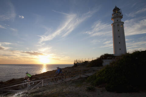 Spanien, Andalusien, Mann und Frau fahren mit dem Fahrrad am Leuchtturm bei Sonnenuntergang, lizenzfreies Stockfoto