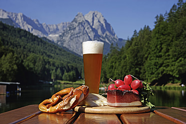 Deutschland, Oberbayern, Bayerische Brotzeit auf Tisch, Berg mit See im Hintergrund - TCF002282