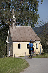 Deutschland, Bayern, Mann radelt über Landstraße, Kapelle im Hintergrund - DSF000332