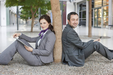 Deutschland, Leipzig, Geschäftsleute sitzen an einem Baum mit digitalem Tablet und Mobiltelefon, lizenzfreies Stockfoto