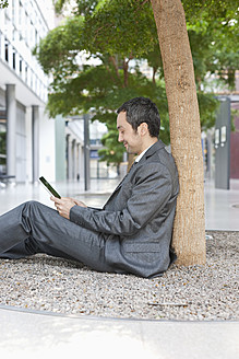 Deutschland, Leipzig, Geschäftsmann sitzt an einem Baum und benutzt ein digitales Tablet - WESTF018572