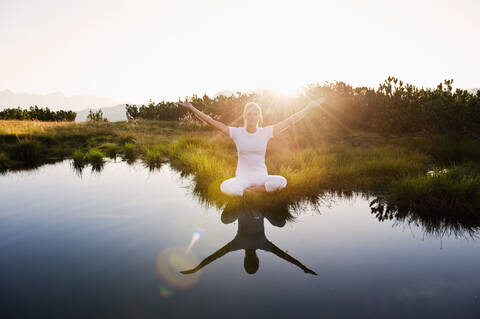 Austria, Salzburg County, Young woman sitting near mountain lake and doing meditation stock photo