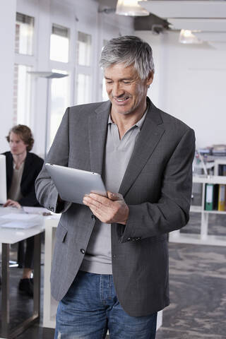 Deutschland, Bayern, München, Älterer Mann mit digitalem Tablet, Kollege arbeitet im Hintergrund, lizenzfreies Stockfoto