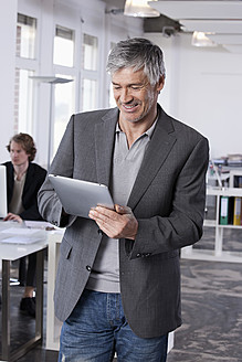 Deutschland, Bayern, München, Älterer Mann mit digitalem Tablet, Kollege arbeitet im Hintergrund - RBYF000020