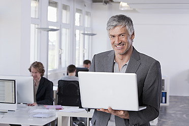 Germany, Bavaria, Munich, Mature man using laptop, colleagues working in background - RBYF000099