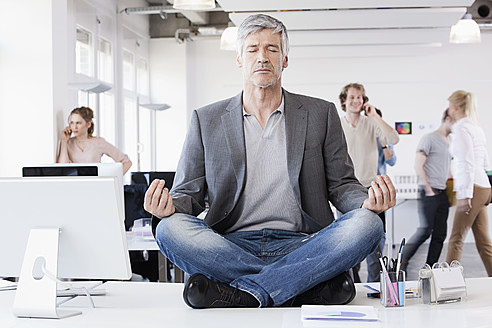 Germany, Bavaria, Munich, Man sitting in lotus position, colleagues in background - RBYF000102