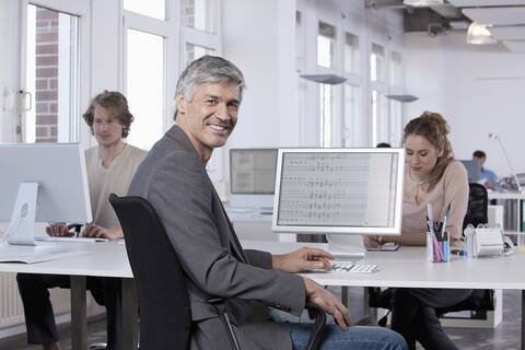 Deutschland, Bayern, München, Älterer Mann lächelnd, Kollegen arbeiten im Hintergrund, lizenzfreies Stockfoto