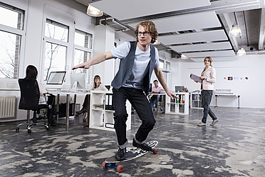 Deutschland, Bayern, München, Mann beim Skateboardfahren im Büro, während Kollegen im Hintergrund arbeiten - RBYF000084