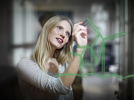 Germany, Cologne, Young woman drawing wind turbine on glass - RHYF000090
