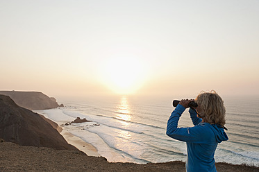 Portugal, Algarve, Sagres, Ältere Frau betrachtet den Strand durch ein Fernglas - MIRF000444