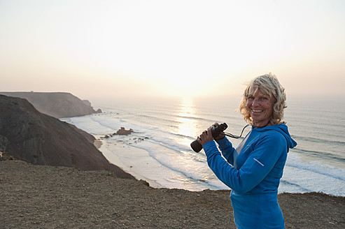 Portugal, Algarve, Sagres, Ältere Frau hält Fernglas am Strand - MIRF000445