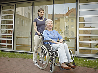 Germany, Cologne, Women pushing senior women in wheelchair outside of nursing home - WESTF018660