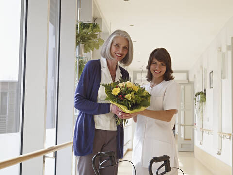 Deutschland, Köln, Seniorin und Pflegerin mit Blumenstrauß im Flur eines Pflegezimmers, lächelnd, Porträt, lizenzfreies Stockfoto