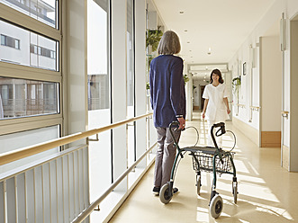 Germany, Cologne, Senior women holding walking frame in corridor, caretaker in background - WESTF018696