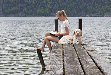 Österreich, Jugendliches Mädchen liest Buch neben Hund auf Steg - WWF002412