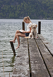 Austria, Teenage girl reading book beside dog on jetty - WWF002383