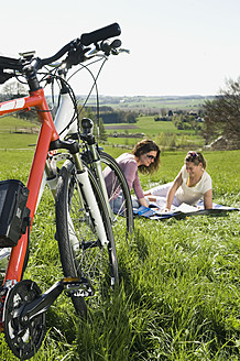 Germany, Bavaria, Mature women sitting on grass and looking at map, electric bicycle in foreground - RNF000935