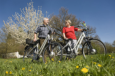 Deutschland, Bayern, Älteres Paar mit Elektrofahrrad auf einer Wiese stehend - RNF000943