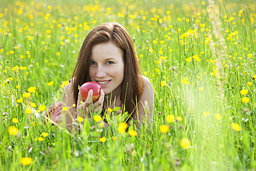 Austria, Young woman lying in field of flowers with apple, smiling, portrait - WWF002424