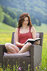 Austria, Young woman sitting on sofa in field of flowers with book - WWF002420