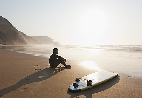 Portugal, Junger Mann am Strand sitzend mit Surfbrett - MIRF000459