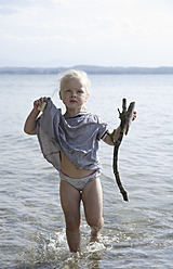 Germany, Bavaria, Girl holding cookie and driftwood, walking in water - TCF002724