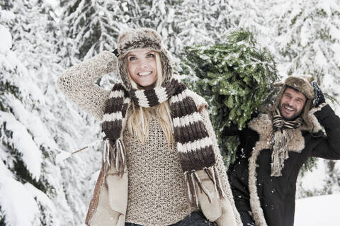 Österreich, Land Salzburg, Mann und Frau gehen durch Schnee, während der Mann einen Weihnachtsbaum trägt, lizenzfreies Stockfoto