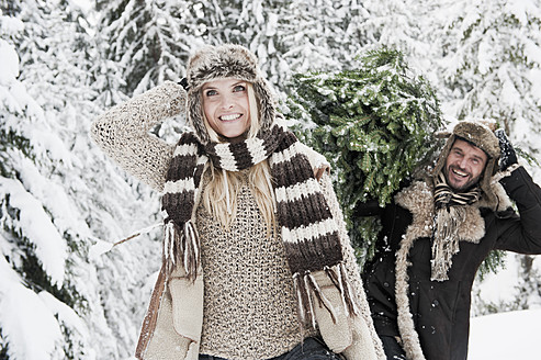 Österreich, Land Salzburg, Mann und Frau gehen durch Schnee, während der Mann einen Weihnachtsbaum trägt - HHF004288