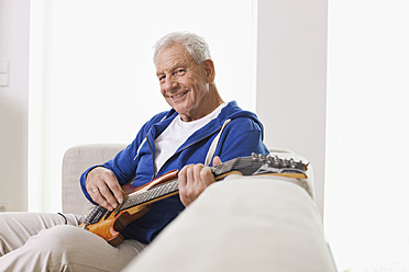 Deutschland, Leipzig, Älterer Mann sitzt auf Sofa und zupft Gitarre - WESTF018878