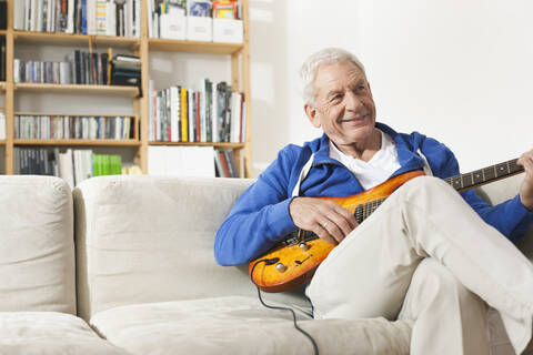 Deutschland, Leipzig, Älterer Mann sitzt auf Sofa und zupft Gitarre, lizenzfreies Stockfoto