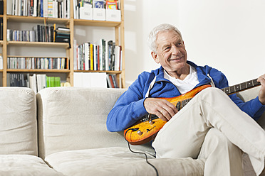 Deutschland, Leipzig, Älterer Mann sitzt auf Sofa und zupft Gitarre - WESTF018879