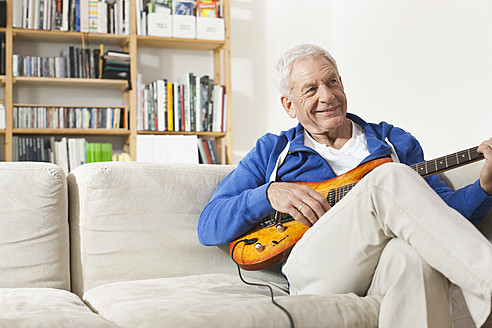 Germany, Leipzig, Senior man sitting on sofa and plucking guitar - WESTF018879