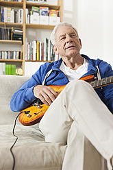 Deutschland, Leipzig, Älterer Mann sitzt auf Sofa und zupft Gitarre - WESTF018880