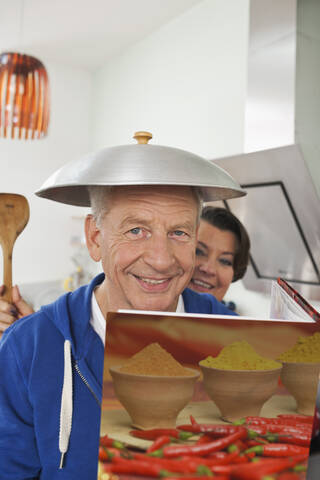 Deutschland, Leipzig, Älterer Mann hält Kochbuch mit Wok-Deckel auf dem Kopf, Frau hält Spachtel im Hintergrund, lizenzfreies Stockfoto