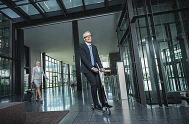 Germany, Stuttgart, Businesswoman with wheeled luggage, man riding scooter at office building - MFPF000212