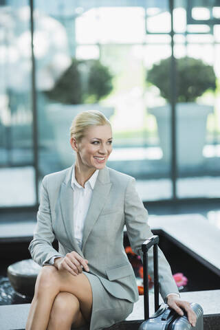 Deutschland, Stuttgart, Geschäftsfrau sitzend mit Gepäck auf Rädern, lächelnd, lizenzfreies Stockfoto