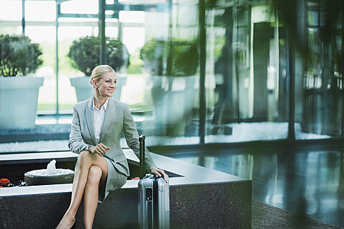 Germany, Stuttgart, Businesswoman sitting with wheeled luggage, smiling - MFPF000217