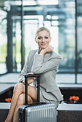 Germany, Stuttgart, Businesswoman sitting with wheeled luggage, smiling, portrait - MFPF000219